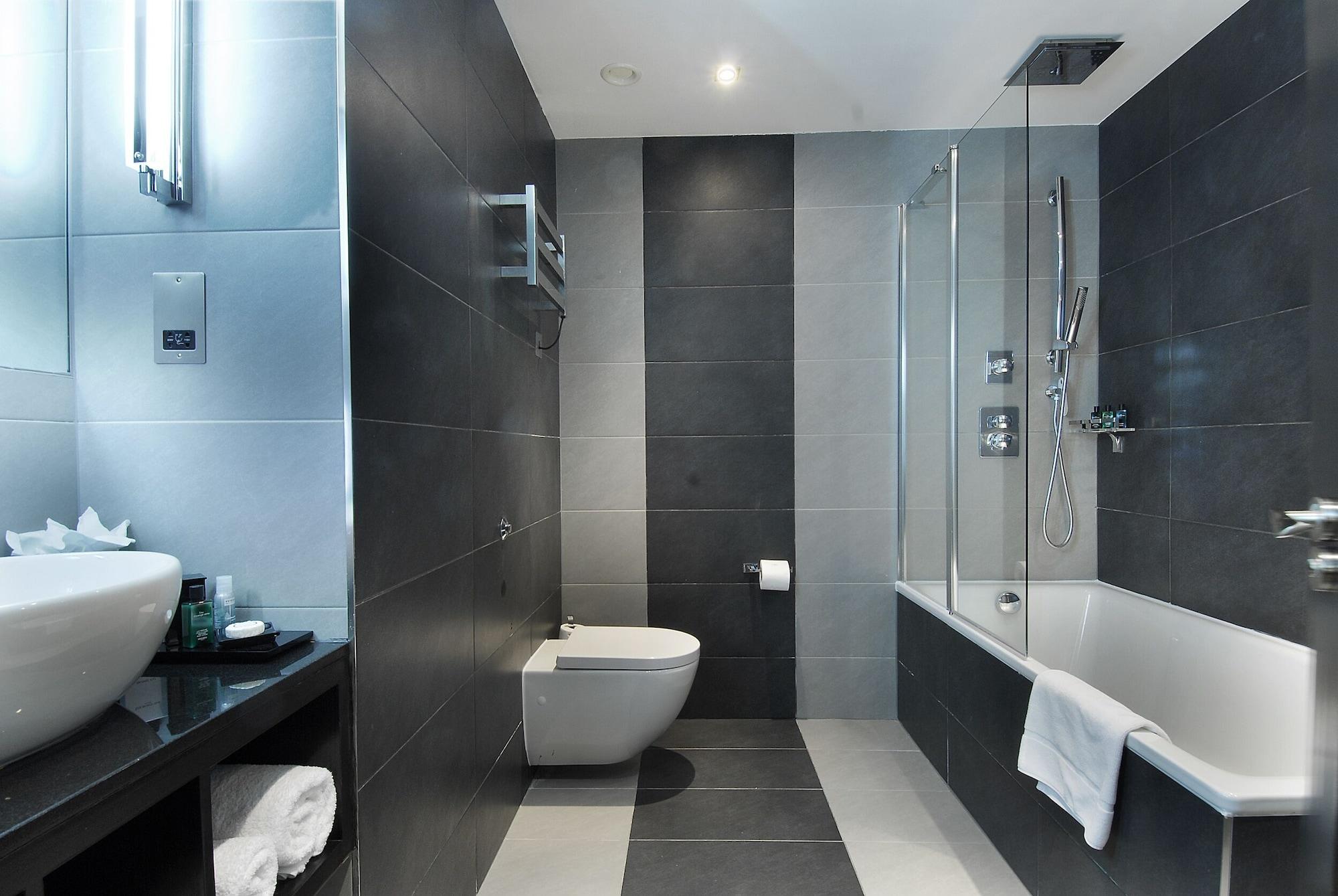 Montcalm Brewery Townhouse Hotell London Exteriör bild The photo shows a modern bathroom featuring a sleek design. The walls are tiled in a combination of dark gray and light gray, creating a contemporary look. On the left, there is a countertop with a basin and some toiletries placed on it. The middle s