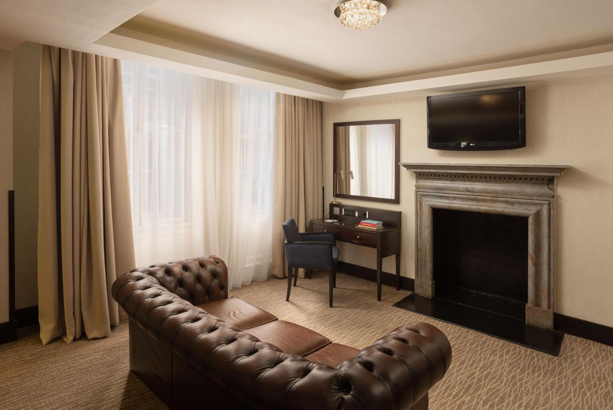 Montcalm Brewery Townhouse Hotell London Exteriör bild The photo shows a stylish interior of a room, likely a living area or a hotel suite. It features a dark leather chesterfield sofa with tufted upholstery. Opposite the sofa is a small wooden desk with a chair, positioned near a wall-mounted flat-scree
