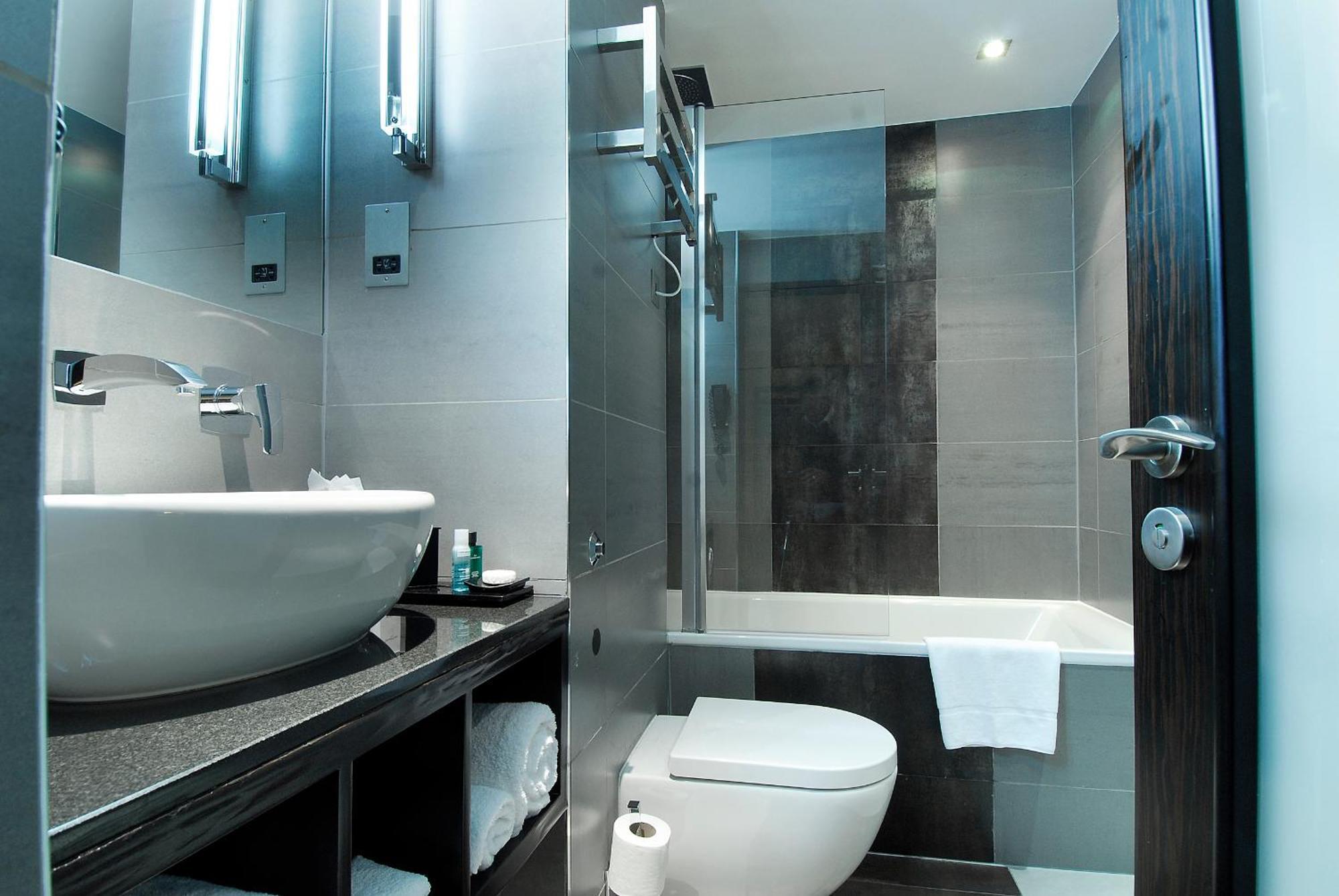 Montcalm Brewery Townhouse Hotell London Exteriör bild The photo shows a modern bathroom with sleek design elements. It features a curved white sink on a dark countertop, with a mirror above that is illuminated by stylish lighting fixtures. Next to the sink, there's a toilet, and on the opposite side, a 