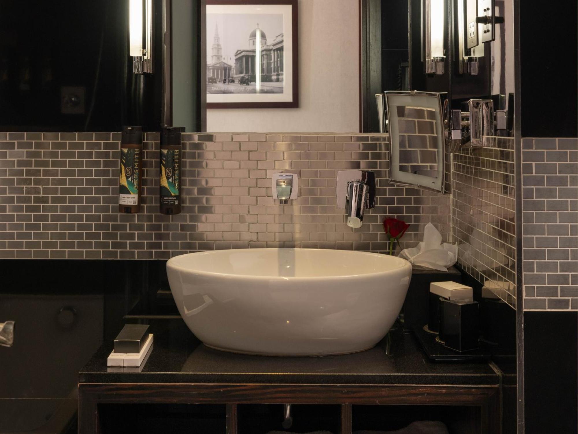 Montcalm Brewery Townhouse Hotell London Exteriör bild The photo shows a modern bathroom with a stylish design. It features a large, round white sink placed on a dark wooden vanity. The walls are adorned with a mix of gray and silver tile, creating a sleek and contemporary look. Above the sink, there is 
