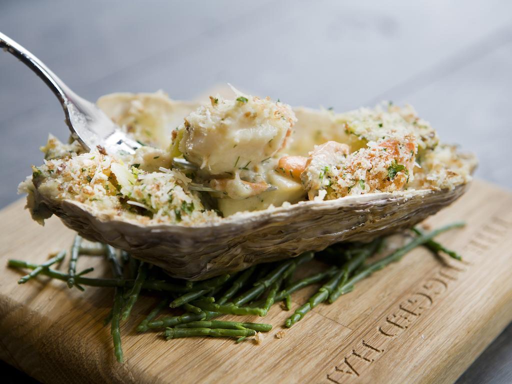 Montcalm Brewery Townhouse Hotell London Exteriör bild The photo shows a large oyster shell filled with a mixture that appears to include seafood, herbs, and breadcrumbs. The dish is presented on a wooden board with some greenery, possibly a type of seaweed or garnishing. A fork is placed beside the oyst