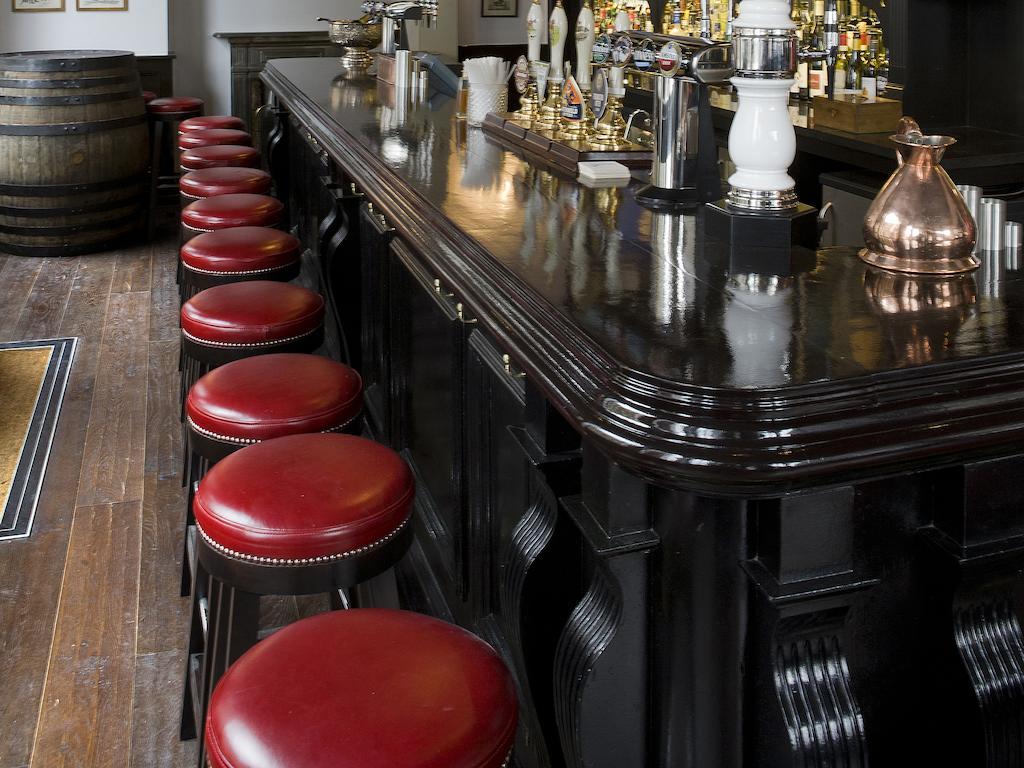 Montcalm Brewery Townhouse Hotell London Exteriör bild The photo shows a bar setting with a long, polished dark wooden bar counter. Attached to the counter are several bar stools with red leather seats. Behind the bar, there are various bottles, suggesting a well-stocked bar. The flooring appears to be w