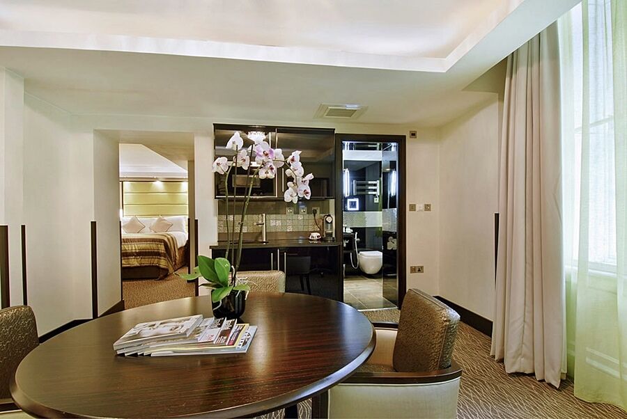 Montcalm Brewery Townhouse Hotell London Exteriör bild The photo shows a stylish hotel room or suite. In the foreground, there is a round dining table with a few magazines on it. Surrounding the table are upholstered chairs. To the right, there are large windows with light-colored curtains, allowing natu